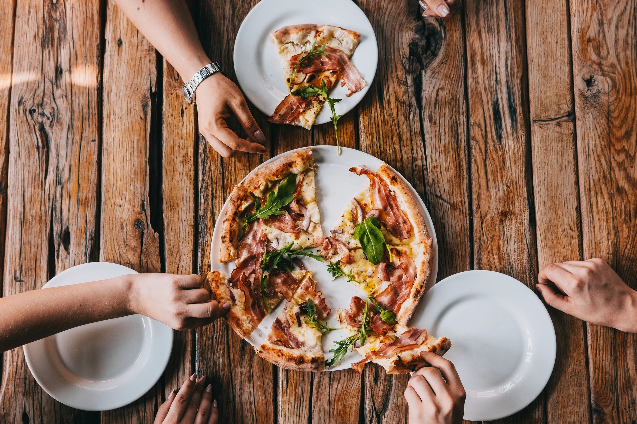 Hands grabbing pizza carbonara on rustic wooden table.